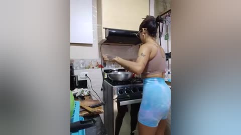 Media: A video of a woman with a medium build, wearing a pink sports bra and blue tie-dye shorts, cooking on a stove in a cluttered kitchen.