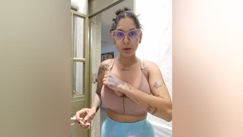 Media: Video of a curvy woman with medium skin tone, large breasts, and tattoos, wearing a pink sports bra and blue leggings, adjusting her bra in a bathroom.