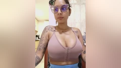 Media: Video of a woman with medium-dark skin, wearing purple glasses and a pink halter top, revealing a tattoo on her chest, sitting in a kitchen with white cabinets.