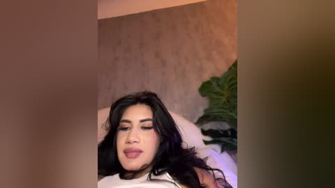 Media: A video of a Latina woman with long, wavy black hair, dark eyebrows, and full lips, lying on a bed with a beige headboard and green plant in the background.