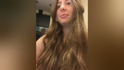 Media: A video of a young woman with long, wavy, light brown hair, standing indoors with a blurred background featuring a kitchen. She has a neutral expression.