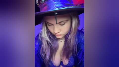 Media: A video of a young woman with long, platinum blonde hair, wearing a shiny, metallic top hat and a revealing blue dress, kneeling with hands clasped in prayer, under purple lighting.