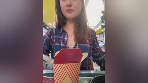 Media: Video of a woman with long brown hair, wearing a plaid shirt, holding a red and yellow striped takeout box. Background shows a dimly lit fast-food restaurant.