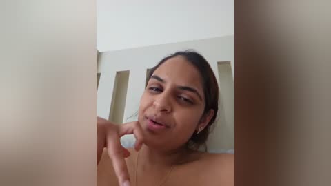 Media: Video of a young woman with medium brown skin, brown hair, and a thin build, making a kissy face, standing in a minimalist room with beige walls and rectangular cutouts.