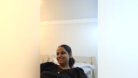 Media: A video of a South Asian woman with medium brown skin, dark hair tied back, wearing a black top and gold necklace, sitting on a hospital bed with white sheets, smiling.