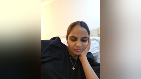 Media: Video of a South Asian woman with medium-brown skin, medium-length black hair, wearing a black t-shirt, resting her chin on her hand in a contemplative pose. Background shows a white wall and a bed with white sheets.