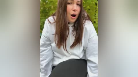 Media: Video of a young woman with long brown hair, wearing a white turtleneck and black pants, seated outdoors with blurred green foliage in the background. She has a surprised or shocked expression.