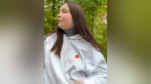 Media: Video of a young woman with long brown hair, wearing a white hoodie with a red logo, standing outdoors in a blurred, green forest background.