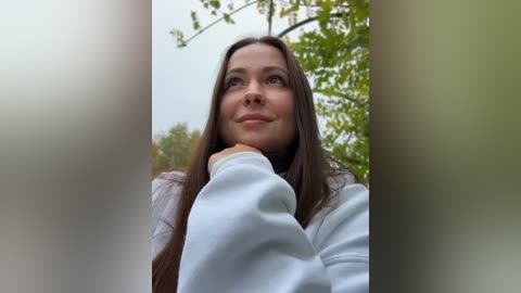 Media: A video of a young woman with long brown hair, fair skin, and a serene expression, wearing a white hoodie, looking upward through blurred green foliage.