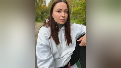 Media: Video of a young woman with light skin, long brown hair, wearing a white hoodie, sitting in a park. Background shows blurred greenery and trees.