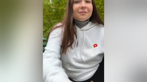 Media: Video of a young woman with long brown hair, wearing a white zip-up hoodie with a red logo, standing in front of greenery.