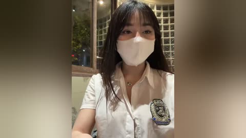 Media: Video of an Asian woman with long black hair, wearing a white uniform with a police badge, a face mask, and a necklace, standing indoors with glass blocks and plants in the background.