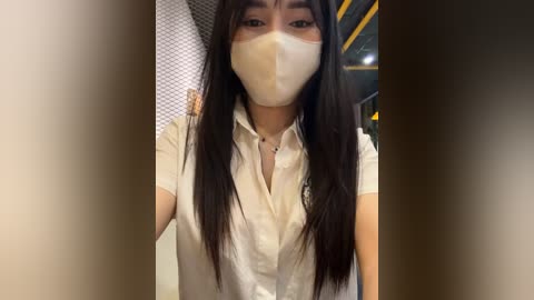 Media: A video of an Asian woman with long black hair, wearing a white blouse, face mask, and a delicate necklace, indoors near a patterned wall.