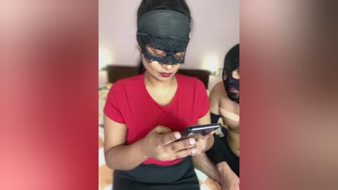 Media: Video of a woman in a red top, black mask, and black skirt, holding a smartphone, with blurred background, emphasizing focus on her face.