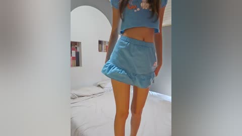 Media: Video of a slender, light-skinned woman with long brown hair wearing a light blue crop top and matching short skirt, standing in a minimalistic, brightly lit bedroom with white walls, a bed, and abstract artwork.