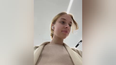 Media: Video of a young Caucasian woman with fair skin and blonde hair, wearing a beige jacket over a beige sweater, looking downwards.