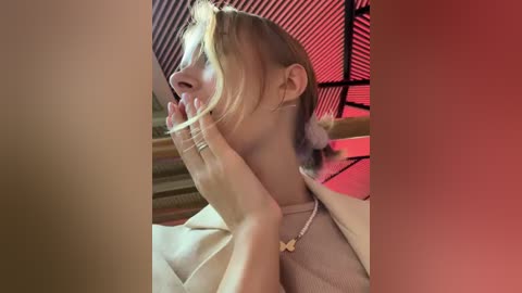 Media: Video of a young Asian woman with light skin, wearing a beige top and a gold necklace, holding her hand to her mouth. She has short, blonde hair. Background features a red-lit, black-framed window.