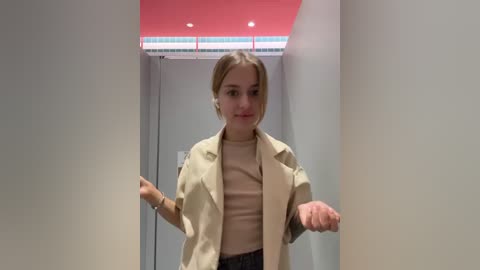 Media: Video of a young woman with blonde hair, wearing a beige blazer and black top, standing in a narrow hallway with red and white ceiling tiles.