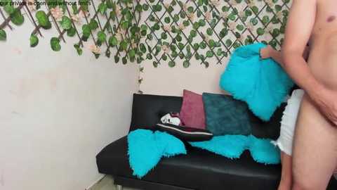 Media: A video of a man wearing white underwear, standing in a room with a black sofa covered in turquoise and teal fur blankets. The walls are adorned with green ivy and floral wallpaper.