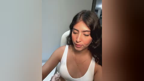 Media: Video of a young woman with long, dark hair, wearing a white tank top, sitting on a white chair in a modern room with light-colored walls.