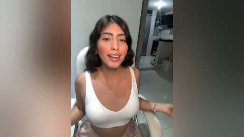 Media: Video of a young woman with dark hair, light skin, wearing a white tank top and pink shorts, sitting on a white chair in a modern, minimalistic kitchen.