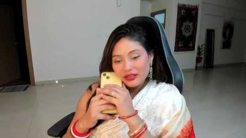 Media: Video of a woman in a white and gold sari, holding a gold phone to her ear, seated indoors with tiled floors and a modern office chair.