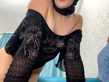 Media: Video of a light-skinned woman with a slender build, wearing an off-shoulder, black lace top revealing her breasts, and a black choker. Background shows a textured wall, teal curtains, and a blue bedspread.