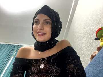 Media: Video of a smiling woman with medium skin tone, wearing a black lace off-shoulder top, black hijab, and a silver necklace. Background features a teal curtain, patterned wallpaper, and a colorful stuffed animal.