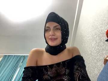 Media: Video of a young woman with fair skin, wearing a black, off-shoulder lace top and a black hijab with sequins, standing indoors against a textured, light-colored wall.