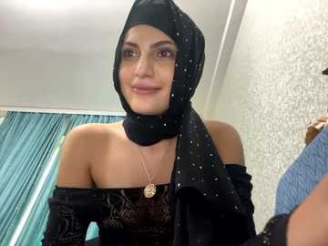 Media: Video of a young woman with light brown skin, wearing a black hijab and off-shoulder black dress, standing indoors near teal curtains and a textured wall.