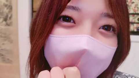 Media: Video of an Asian woman with straight, shoulder-length auburn hair and light skin, wearing a white surgical mask. She has large, expressive brown eyes and is holding her mask. Background features a blurred, patterned wall.