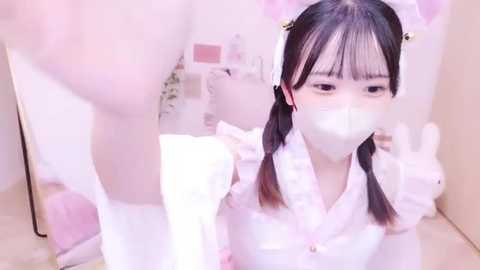 Media: A young East Asian woman with black pigtails and a white face mask, wearing a white maid outfit with frilly details, stands in a pink-themed room, reflecting in a mirror.