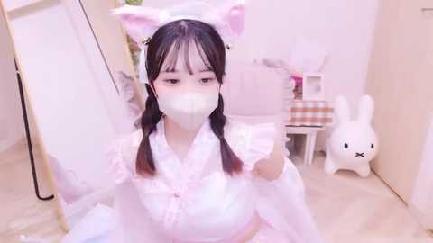 Media: A young Asian woman with fair skin and long black hair styled in twin braids, dressed in a pink bunny costume with fluffy ears, wearing a face mask, takes a selfie in a brightly lit, minimalistic room with a white floor.