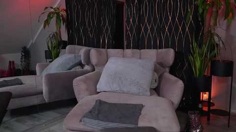 Media: Video of a modern living room with beige sectional sofa, gray pillows, dark patterned wall, potted plants, black lamps, and warm lighting.