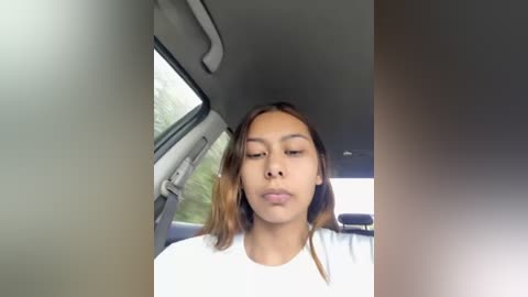 Media: Video of a young woman with medium brown skin, straight brown hair, wearing a white t-shirt, sitting in a car with light grey interior and a window showing greenery outside.