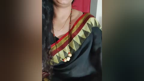 Media: Video of a woman in a black saree with gold and red geometric patterns, wearing a gold necklace with red and white beads. Her long, black hair partially obscures her face. Background is blurred, focusing on the saree.