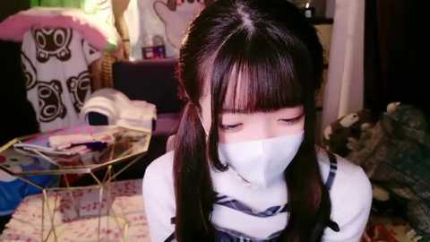 Media: Video of a young Asian girl with long black hair and a face mask, sitting on a colorful rug in a cluttered room with a pink bear and a glass table.