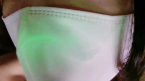 Media: Video of a person's face, partially covered by a white mask with a green filter, showing the mask's elastic bands and fabric texture.