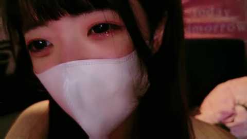 Media: A close-up video of a young Asian woman with long black hair and brown eyes, wearing a white surgical mask, taken indoors with a blurred background.