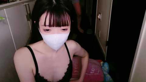 Media: A video shows an Asian woman with long black hair, wearing a white mask, black lace bra, and a pink blanket in a dimly lit, cramped room.