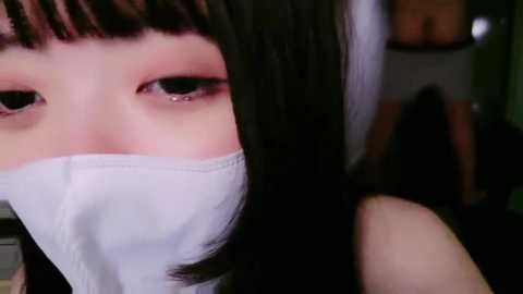 Media: Video of a young woman with pale skin, black hair, and dark eyes, wearing a white surgical mask, in a dimly lit room with blurred furniture in the background.