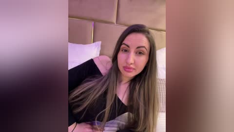 Media: Video of a young woman with long, straight, dark hair, wearing a black top, lying on a bed with white pillows and beige headboard, in a cozy, well-lit bedroom.