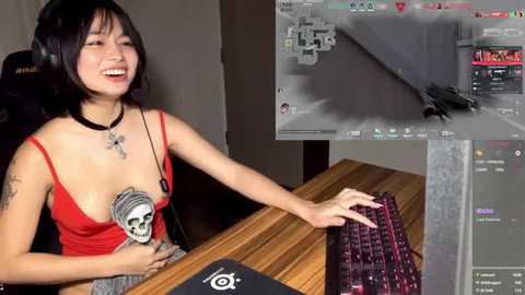 Media: Video of an Asian woman with shoulder-length black hair, wearing a red spaghetti-strap top, laughing while playing a video game on a wooden desk. She has a skull tattoo on her chest.