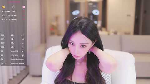 Media: A video of an Asian woman with long black hair, wearing a black off-shoulder top, sitting in a modern living room, hands behind her head. The background features blurred furniture and soft lighting.