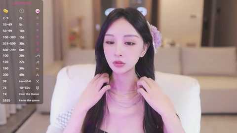 Media: Video of a young Asian woman with long black hair, wearing a floral headband and a pink top, adjusting her necklace in a modern, dimly lit living room.