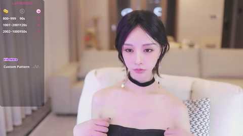 Media: Video of an East Asian woman with pale skin and black hair, wearing a black choker and a black off-shoulder top, sitting on a white couch. The background is a modern, dimly-lit living room.