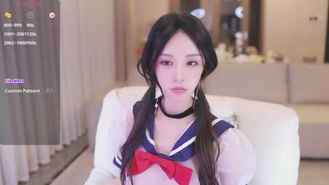 Media: Video of an Asian woman with long black hair in pigtails, wearing a Japanese schoolgirl outfit with a blue sailor collar and red bow. Background features a modern, beige-toned room with curtains and furniture.