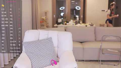 Media: Video of a modern living room with a white sofa, black-patterned cushions, and a transparent chair. A woman in a red dress stands near a table with a drink.