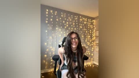 Media: Video of a young woman with long, wavy brown hair, wearing glasses, and a plaid shirt, seated on a black office chair in a room with a beige wall and a string of golden fairy lights.