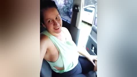 Media: A video shows a smiling woman with dark hair and fair skin, wearing a light green tank top, sitting in a car. The background features a parked white car and greenery outside.